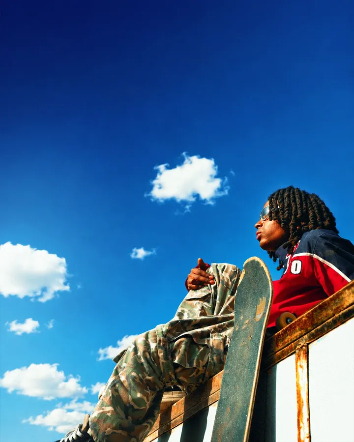 Low-angle frame seated on a metal edge holding a skateboard against blue sky and clouds.