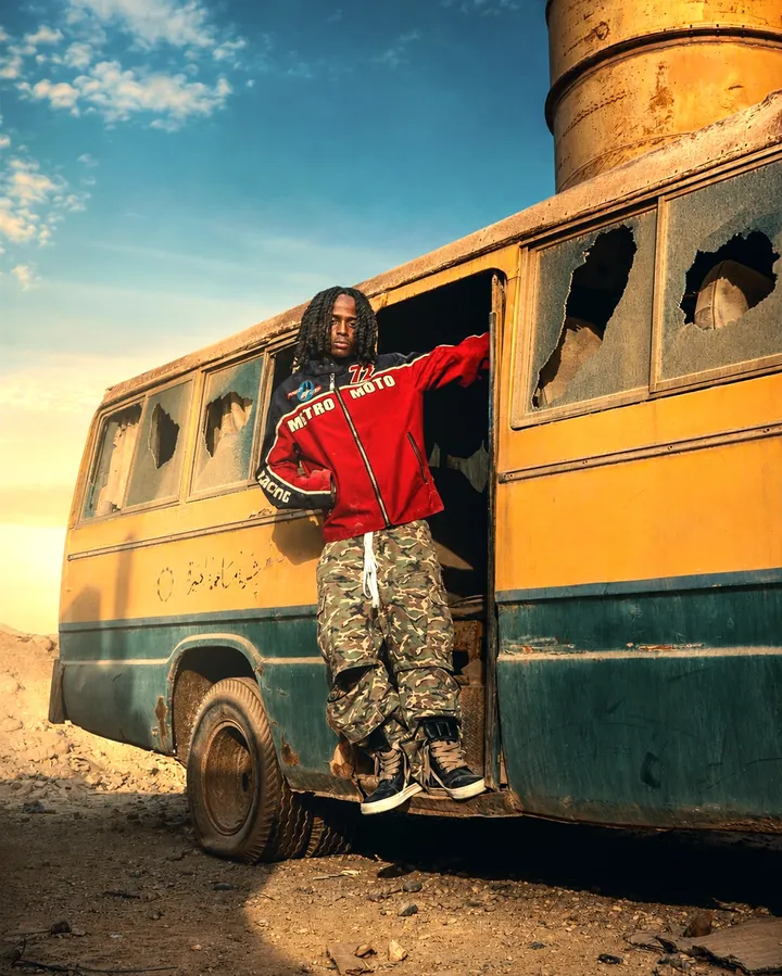 Leaning out of a derelict bus doorway in a red jacket and camo trousers on open dirt ground.