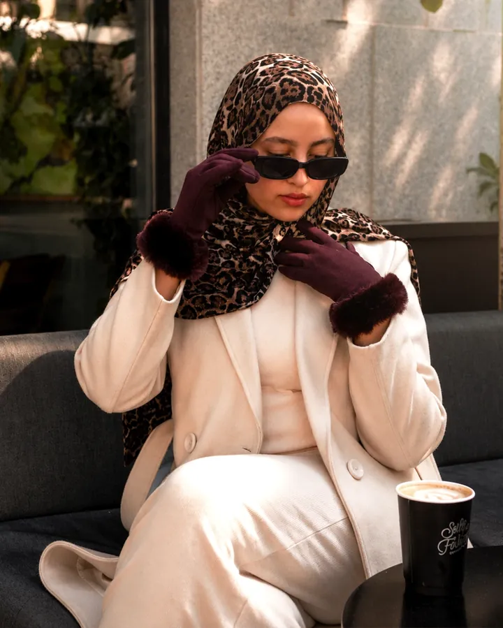 A gloved hand raised near the face, paired with a leopard-print scarf and cream coat.