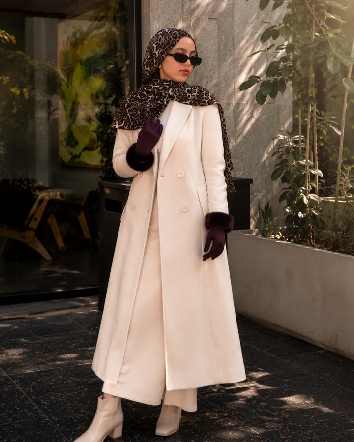 Standing full-length in a long cream coat and burgundy gloves against a naturally lit wall.