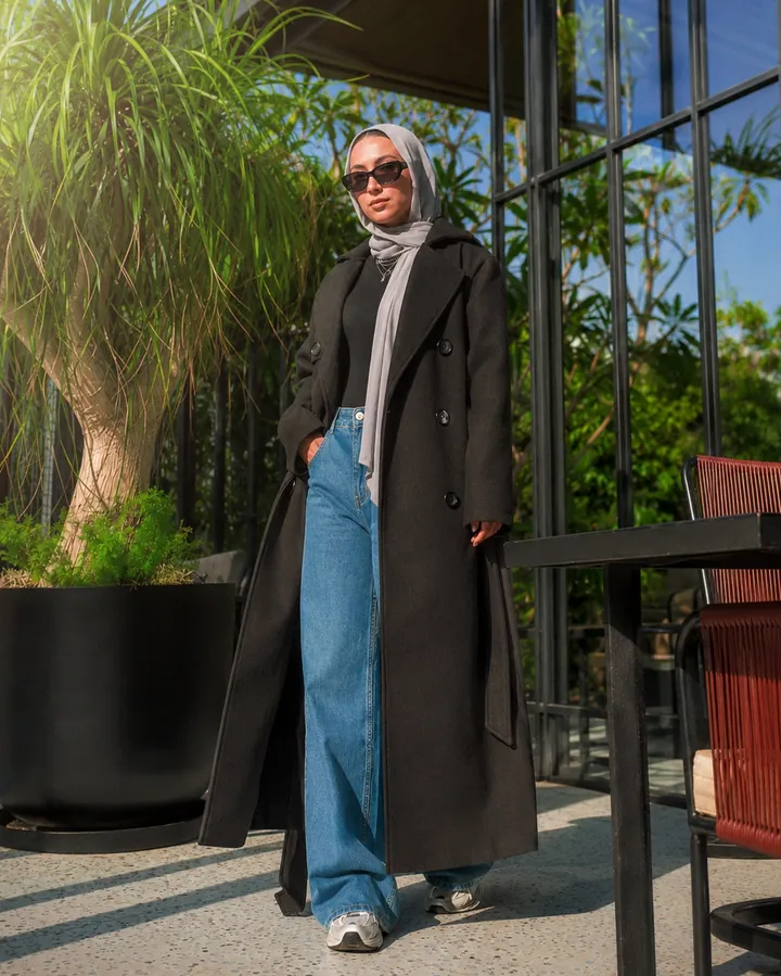 Full-length look of a long black coat over wide jeans in bright daylight against a glass facade.