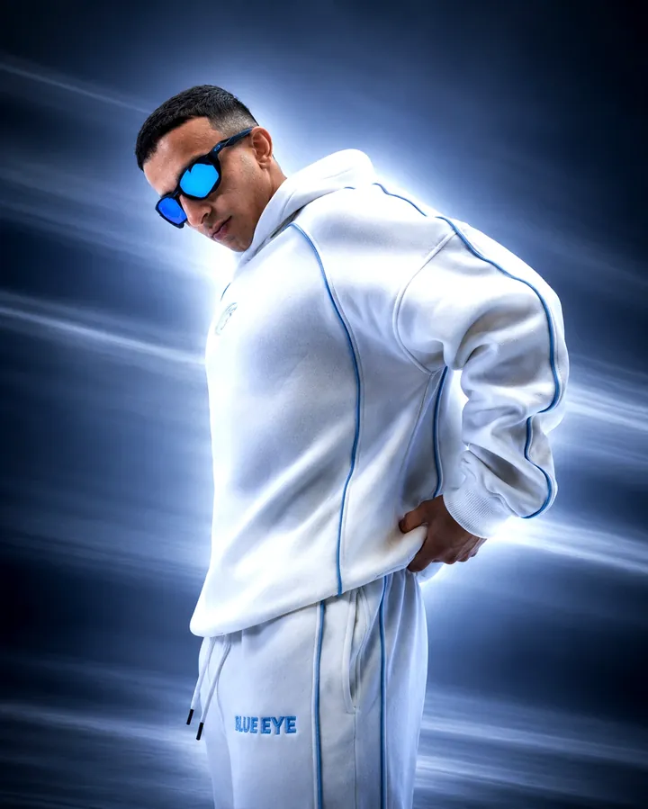 A side turn in a white hoodie with blue piping and mirrored blue sunglasses under studio light.