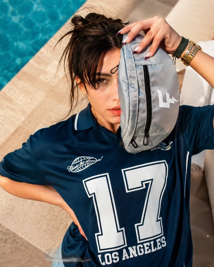 Overhead frame of a woman in a navy sports jersey holding a grey belt bag.