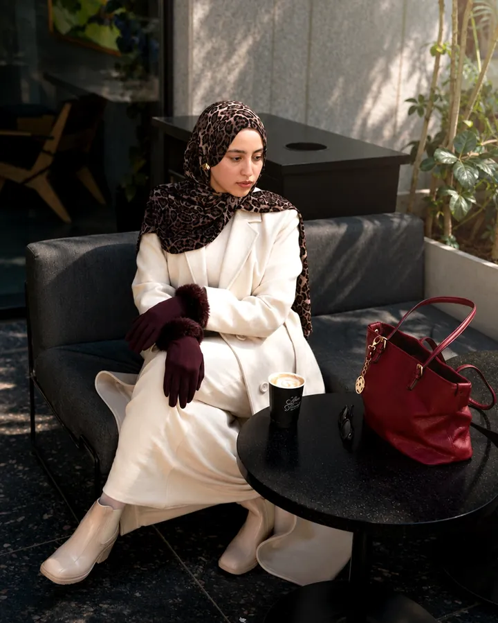 Seated on a café sofa with a coffee cup, a red bag and a long cream coat.